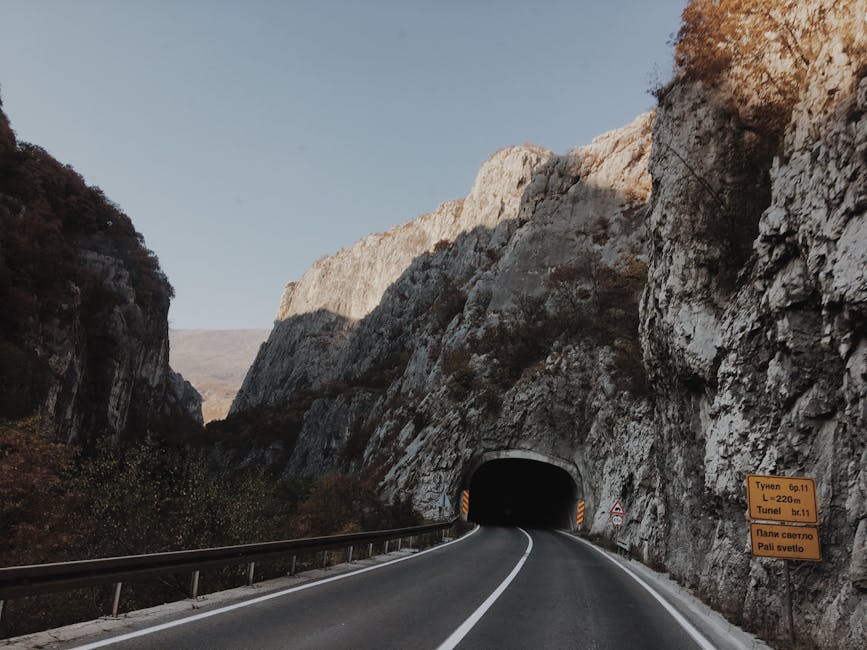 A picturesque road winds through rocky mountains, entering a tunnel under clear skies.